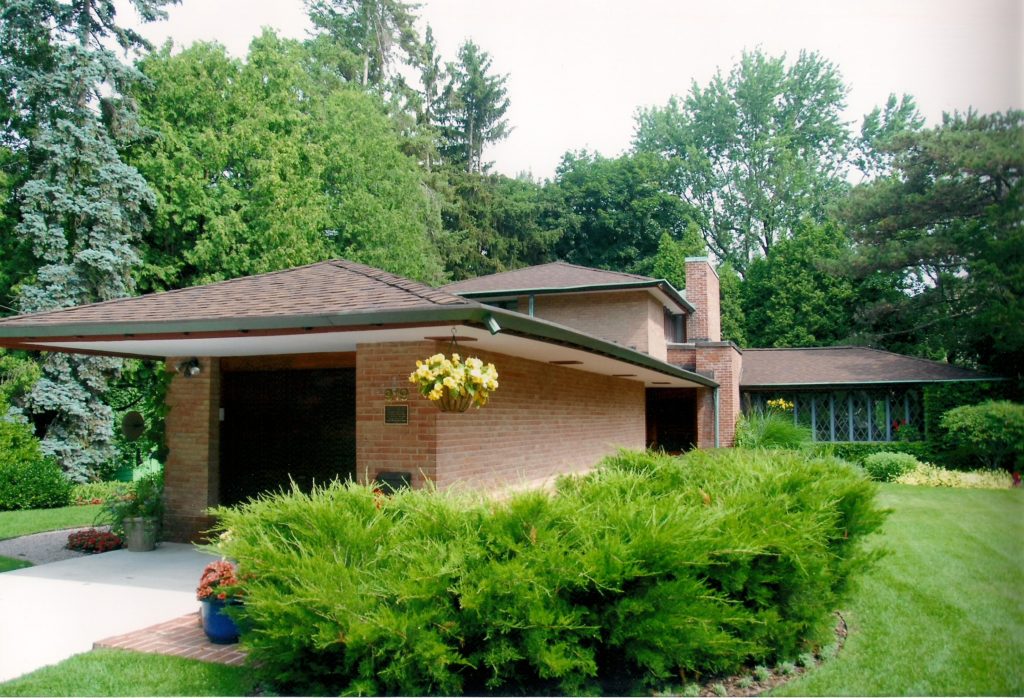 Exterior of a midcentury modern brick home surrounded by bushes and with yellow flowers hanging from the corner of the roof
